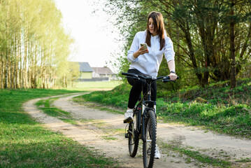 Young woman riding a bicycle and holding a phone in her other hand in the forest at sunset.