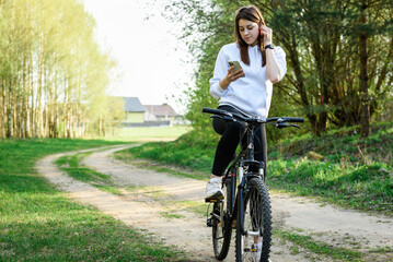 Fototapeta premium Young woman riding a bicycle and holding a phone in her other hand in the forest at sunset.