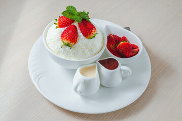 Bowl of shaved ice dessert topped with fresh strawberries and mint, served with strawberry sauce and condensed milk on a white plate, Sweet Dessert.