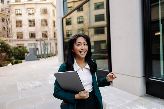 Businesswoman walking in city street holding a laptop and smiling - Powered by Adobe
