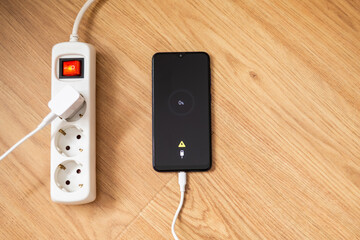 White extension cord plugged into wall socket powers smartphone displaying low battery warning on wooden table, emphasizing the importance of accessible power sources in modern life