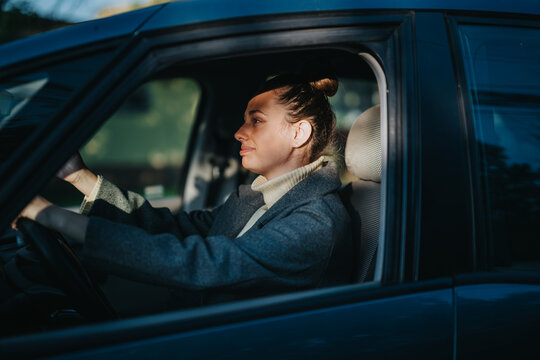 A woman drives a car through the city with confidence and focus. The warm sunlight casts a gentle glow, highlighting her serene expression and creating a calm atmosphere in the vehicle.