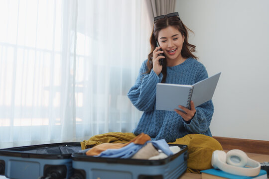 Happy young woman packing clothes into a suitcase, chatting on her smartphone, and reviewing notes from her notebook while eagerly preparing for an exciting trip ahead