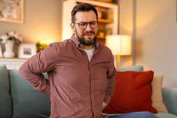 Young man experiencing sharp lower back pain after long hours of sitting, holding his back with a strained expression. Concept of spinal discomfort related to sedentary work and modern lifestyle.