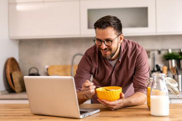 A man in glasses enjoys breakfast while working on his laptop in a modern kitchen. He balances productivity and healthy eating, starting his day with cereal and milk in a cozy, homey atmosphere.