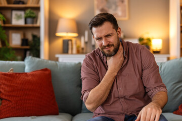 Fototapeta premium Man sitting on a sofa, holding his throat in discomfort, feeling a sore throat. He experiences a moment of pain while resting at home, reflecting the struggle of dealing with a cold or illness.