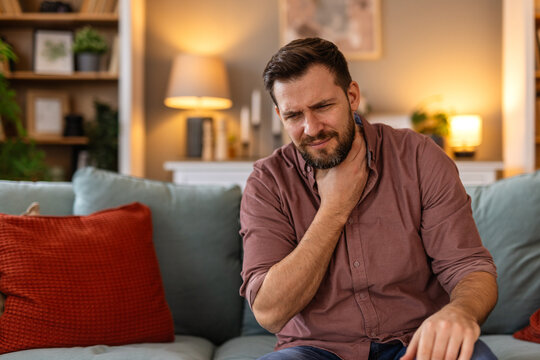 Man sitting on a sofa, holding his throat in discomfort, feeling a sore throat. He experiences a moment of pain while resting at home, reflecting the struggle of dealing with a cold or illness.