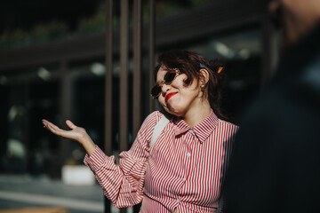 A woman in sunglasses and a striped shirt shrugs with a playful, carefree expression. She conveys a...