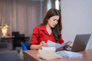 Young Asian businesswoman having problems with domestic bills, checking financial documents, using laptop and calculating domestic expenses sitting at home office desk