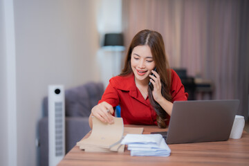Obraz premium Young Asian businesswoman talking on mobile phone and checking documents while working at home office with laptop computer, happy and smiling female freelancer working from home