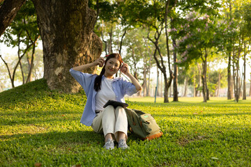 A woman is sitting on the grass in a park, listening to music. She is wearing headphones and has a book in her lap. Concept of relaxation and leisure
