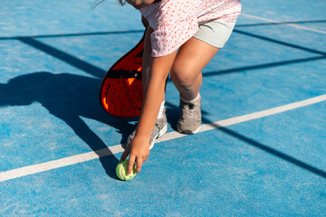 Child picking up tennis ball on sunlit court, ready to play.