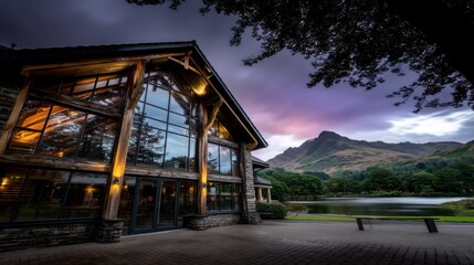 Modern stone-faced building with glass windows and wooden beams near lake and mountains at twilight
