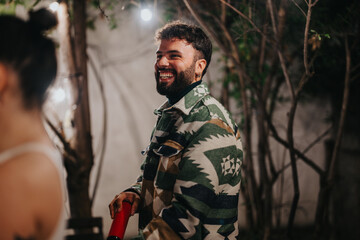 Obraz premium A man in a patterned jacket smiling during an outdoor nighttime party setting. Fairy lights and natural surroundings create a joyful and celebratory atmosphere, capturing warmth and camaraderie.