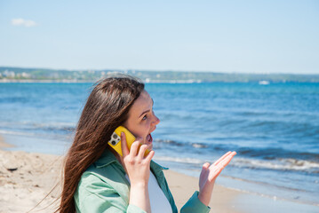 Young caucasian female with long brown hair engaged in animated phone conversation on sunny beach, expressing surprise with blue ocean backdrop. Digital nomad lifestyle. Remote work