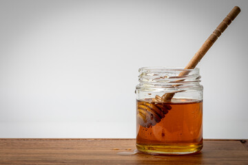 Close up shot of honey dipper immersed in a jar of honey with a white background and wooden board