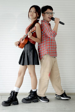 Asian girl holding ukulele and boy holding a microphone while they standing in white room. Elementary school chidden playing music and singing together. Kid with music and instrument.