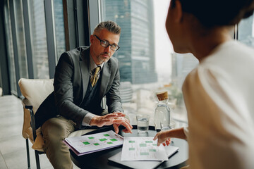 In a highrise office, two business professionals engage in an insightful discussion over various project charts