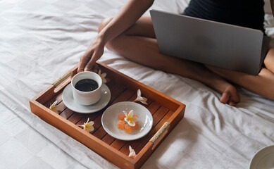 A young woman sitting on the bed and get some coffee while she working on computer.
