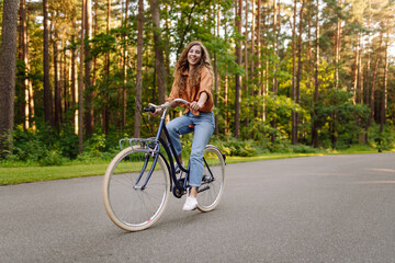 A joyful woman with curly hair rides a bicycle along a path in a sunny park. Young woman having fun...