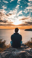 Fototapeta premium A man sits alone on a clifftop overlooking a stunning sunset on the ocean.