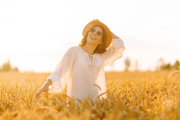 Portrait of happy woman in hat and sunglasses having fun in golden agricultural field. Tourist enjoying sunset in wheat, feeling freedom. Style and beauty concept, relaxation.