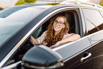 Beautiful woman driving a car, enjoying the ride. Young female driver in glasses at sunset....