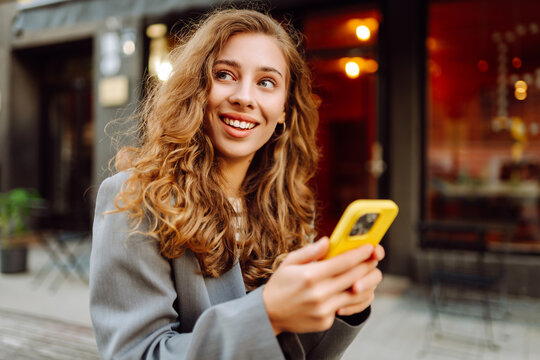 Stylish woman with curly hair smiling with a phone in her hands. Young tourist enjoying a walk around the city, blogging from her phone. Walking concept.