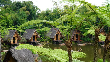 Traditional wooden huts by a serene lake, surrounded by lush greenery and tall ferns at Dusun Bambu Bandung.