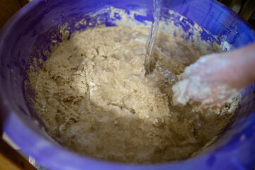 Hands kneading dough in a large plastic container during the process of preparing homemade bread. The dough is sticky and textured, showing active mixing.