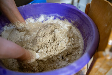 Hands kneading dough in a large plastic container during the process of preparing homemade bread. The dough is sticky and textured, showing active mixing.