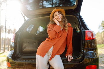 Joyful woman sits in the trunk of a car at sunset in the forest. Traveler enjoys nature, feels...