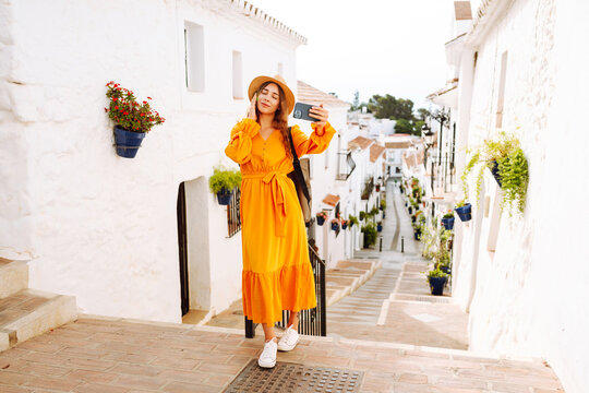 Cheerful tourist in dress and with phone walking along sunny street with white houses. Beautiful woman in hat taking selfie and enjoying travel. Blogging and tourism concept.
