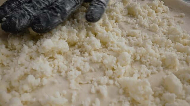 Professional baker wearing black protective gloves generously spreading crumbled cheese over thin rolled dough, preparing artisan pizza with precise culinary technique and attention to detail