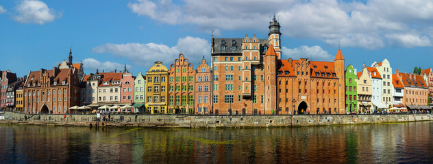 Panoramic view of colorful historic buildings along the Motlawa River in Gdansk, Poland. Features vibrant architecture with a backdrop of blue sky and scattered clouds