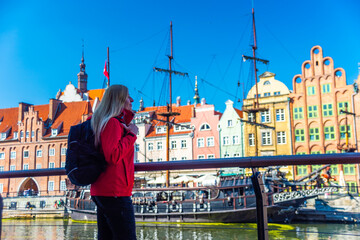 Tourist in red jacket with backpack walks along Gdansk waterfront. Behind her are historical buildings with vibrant facades and a traditional sailing ship at the harbor.