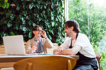 Cafe owner and financial adviser share ideas with smiles in a vibrant cafe space. They review plans at a laptop, symbolizing positive collaboration, small business support, and financial growth.