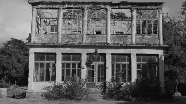 Crumbling facade of a two-story neoclassical building with broken arched windows and weathered pillars, evoking decay and abandonment in monochrome. Documentary shot. History. Background