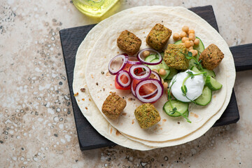 Wheat wraps with falafel, vegetables and yogurt on a black wooden serving board, horizontal shot on a beige granite background, elevated view