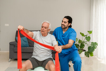 Elderly man on fitness ball doing physiotherapy exercises using a stretch band at home with physiotherapist