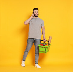 Man with shopping basket full of products talking on smartphone against orange background