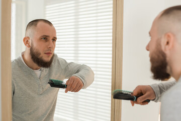 Baldness problem. Worried man brushing hair near mirror at home