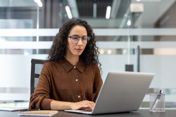 A focused professional woman with curly hair working on a laptop in a modern office setting. She is...