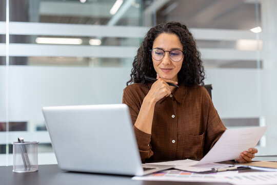 A woman with glasses, working in an office setting, thoughtfully examines documents while using a laptop.