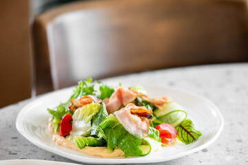 Fresh salad with crispy bacon, cherry tomatoes, cucumber slices, and mixed greens served on a white plate with creamy dressing. The dish is presented on a speckled countertop with a blurred background
