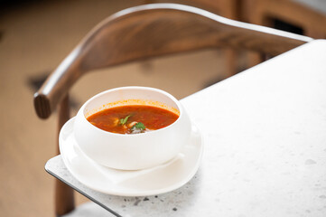 A bowl of fresh tomato soup with cilantro garnish on a white plate, placed on a marble table in a cozy dining setting. Warm and inviting atmosphere, perfect for a comforting meal.