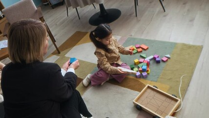 View from above of female Caucasian social worker communicating with little girl playing blocks on floor during appointment