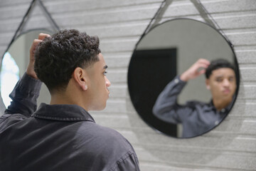 Dominican teenage boy wearing a shirt and earrings fixes his taper fade haircut in a shopping...