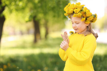 Cute little girl with beautiful flower wearing wreath of dandelions in park, space for text