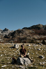 Young woman sitting on a rock with dog in a Montenegro mountain landscape, looking up at the sky and smiling. Peaceful autumn hike scene, connection with nature and pet.